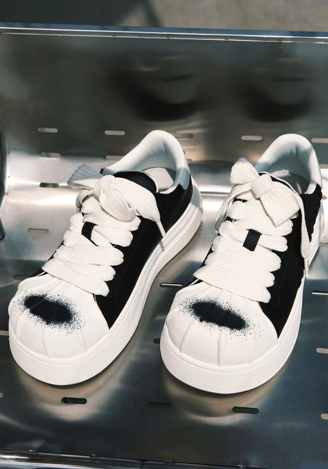 Overhead flat lay of black and white shell toe platform sneakers on a metal shelf, showing broad white laces, thick midsoles, and black spray paint toe accents