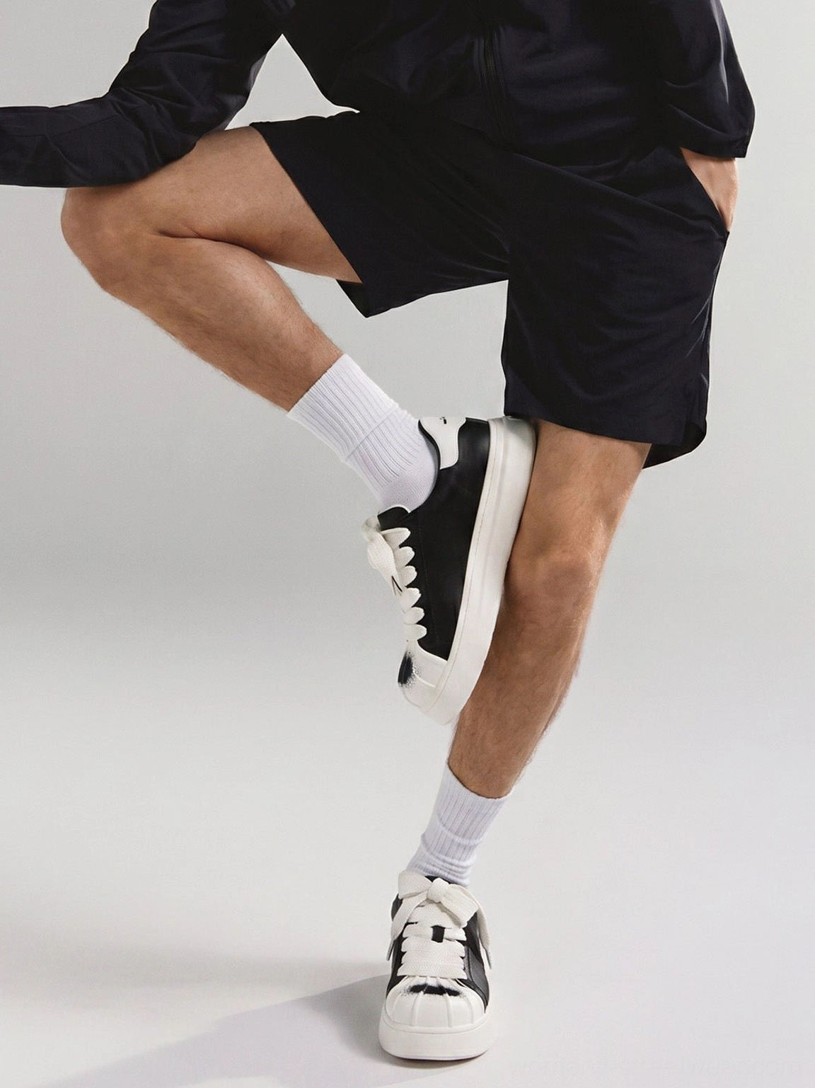 Low-angle view of black and white shell toe platform sneakers worn with black athletic shorts and white crew socks in a coordinated streetwear outfit