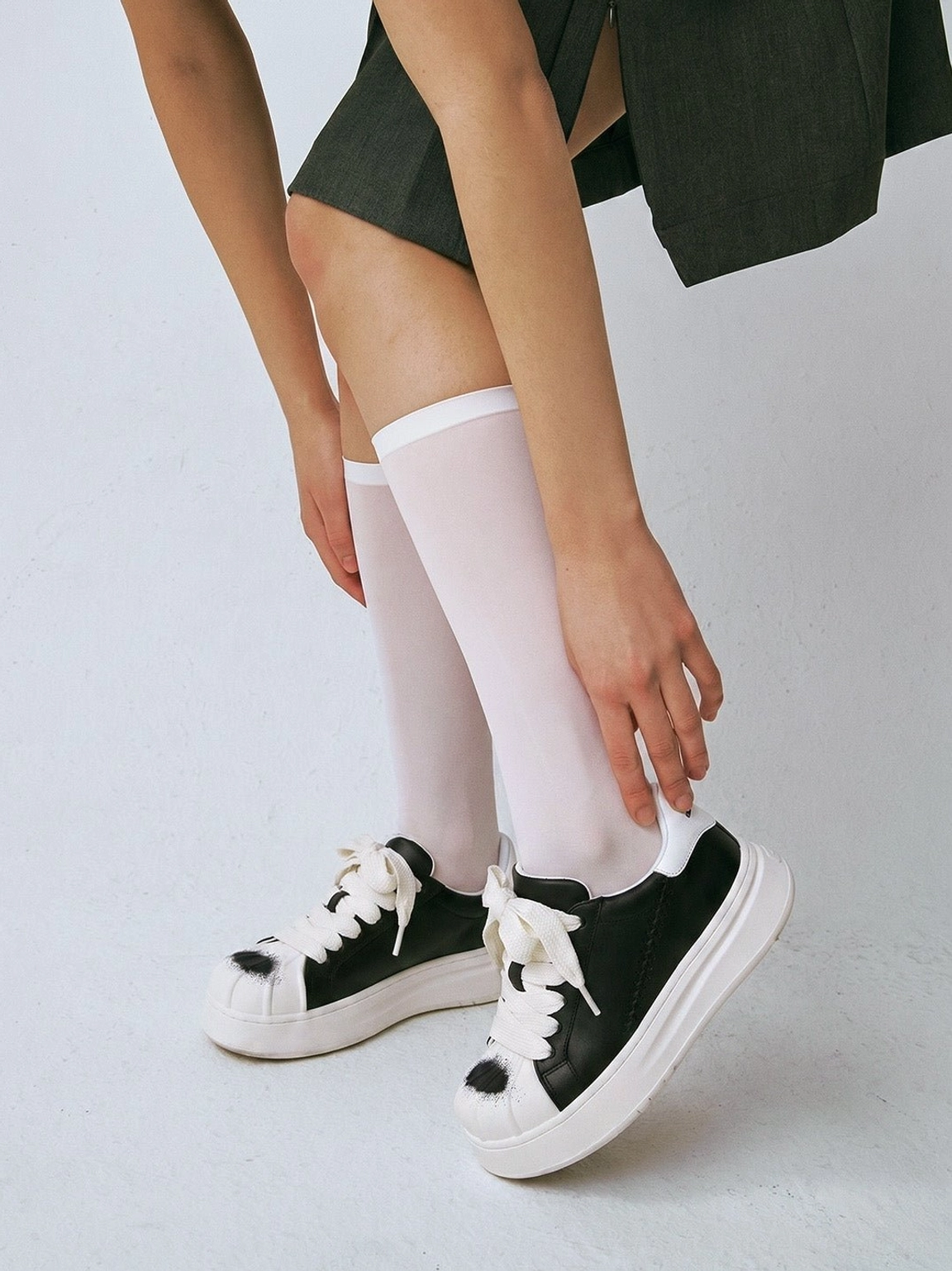 Close-up of black and white shell toe platform derby sneakers worn with long white socks and a dark green mini skirt, featuring black spray accents on the toe caps
