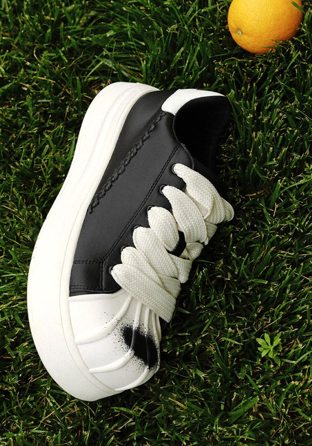 Black and white shell toe platform derby sneaker shown outdoors on grass with wide white laces, black spray toe detail, and woven heel stitching