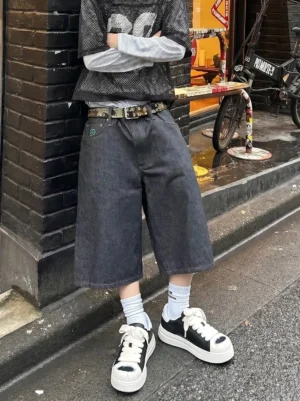 Unisex black and white shell toe platform sneakers styled with cropped wide denim shorts, white crew socks, and layered urban streetwear beside a brick building