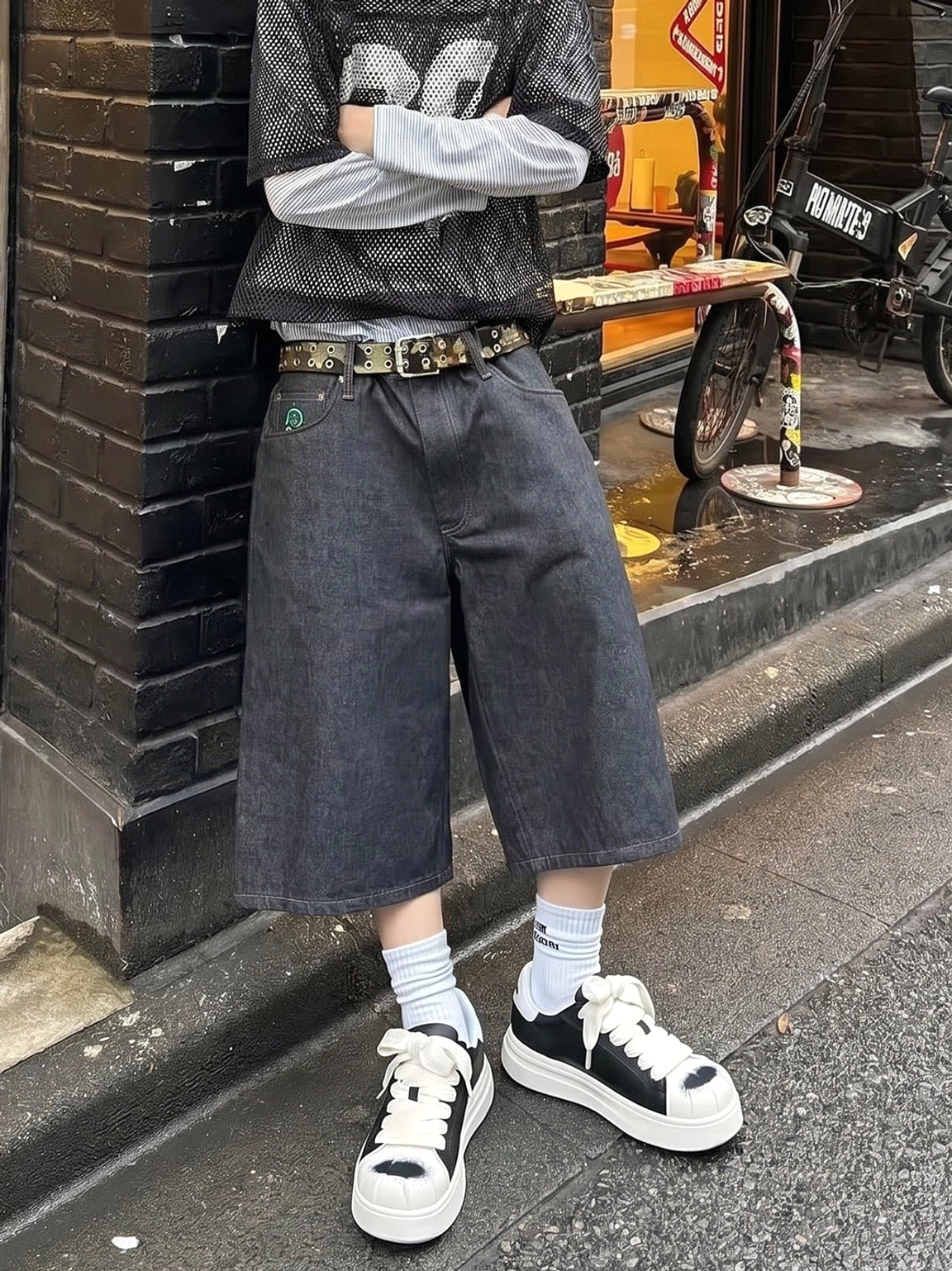 Unisex black and white shell toe platform sneakers styled with cropped wide denim shorts, white crew socks, and layered urban streetwear beside a brick building