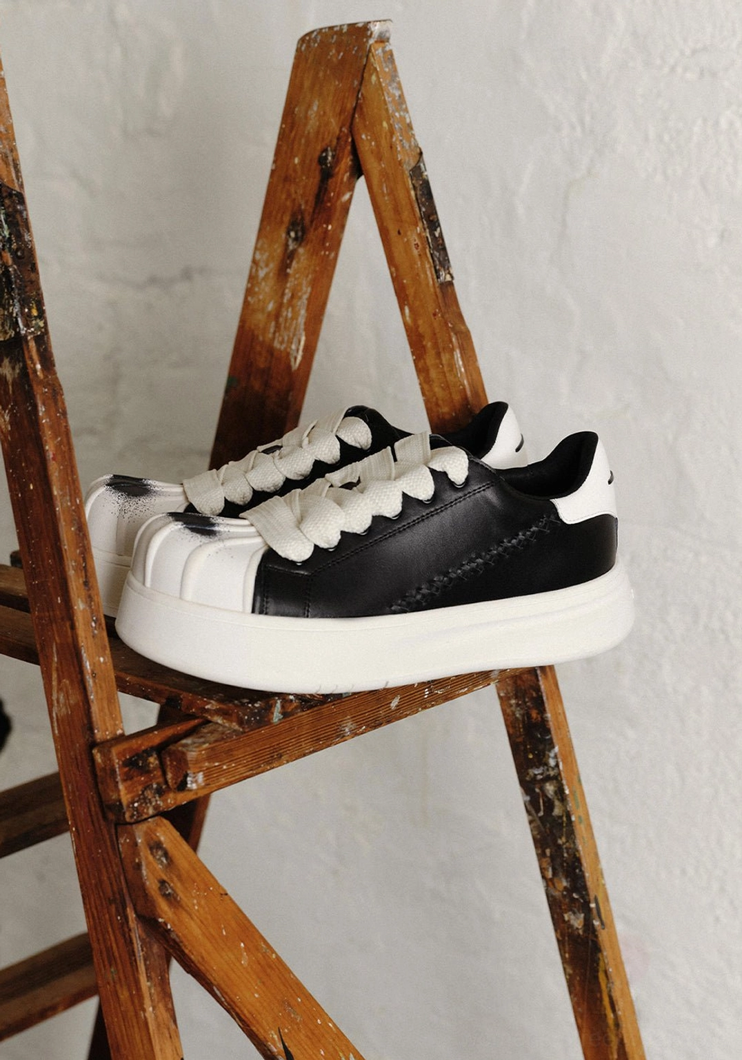 Black and white shell toe platform derby sneaker pair displayed on a wooden ladder with oversized white flat laces, black upper panels, and raised white soles