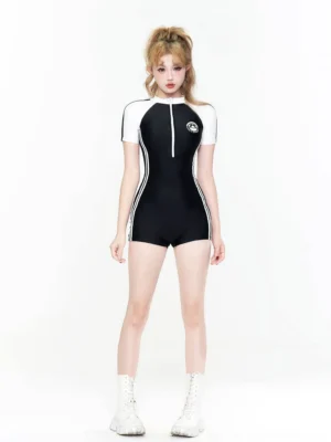 Front full-body view of a black and white short-sleeve zip-front surf romper swimsuit with contrasting raglan sleeves, white side racing stripes, chest patch, and fitted boxer-style swim shorts