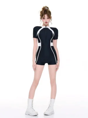 Front full-body view of a black and white short-sleeve zip-up surf swimsuit with high neckline, white contrast piping, boyleg shorts, and athletic streetwear silhouette