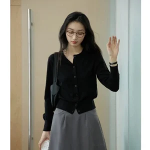 Black women's cropped crew neck button-up cardigan paired with a grey midi skirt and shoulder bag in a modern indoor foyer