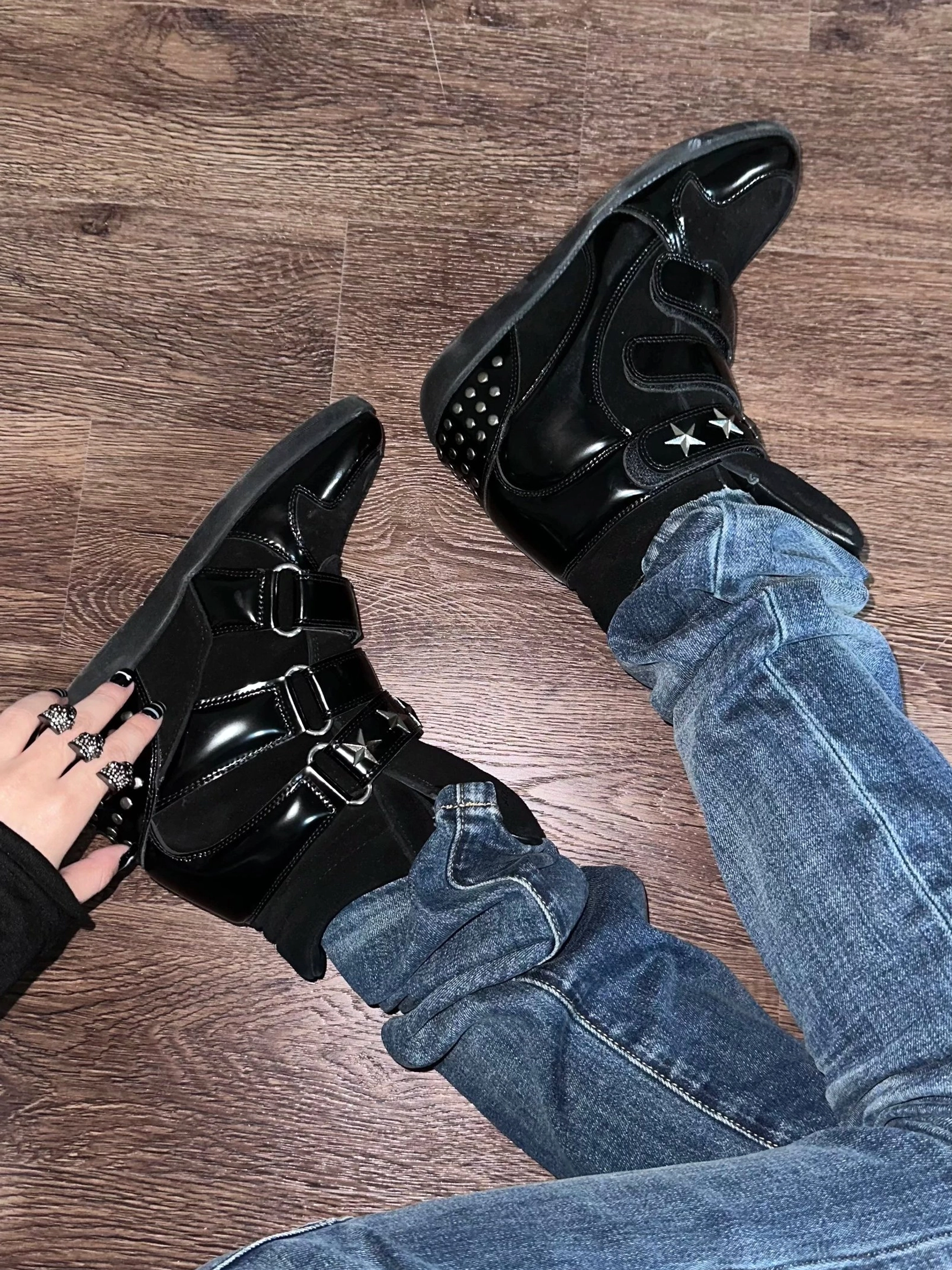 Black women’s hidden wedge high-top sneaker boots worn with blue jeans, featuring suede and patent-effect panels, triple hook-and-loop straps, star metal accents, and a studded heel