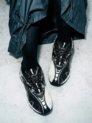 Black and off-white techwear runner sneakers worn with black stockings and a long dark coat on a textured concrete floor