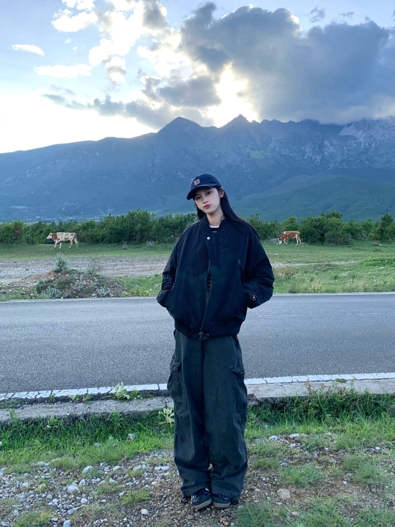 Black zip-up bomber jacket styled with loose cargo trousers and baseball cap beside a mountain highway