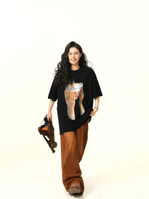 Black oversized artistic graphic tee on a woman with curly hair, featuring a blindfolded face and hands print styled with brown wide-leg pants