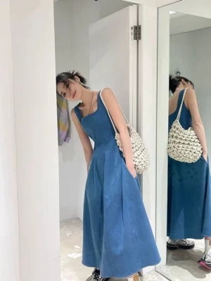 Women's blue denim square-neck maxi dress styled with a crocheted white tote bag in an indoor doorway mirror view