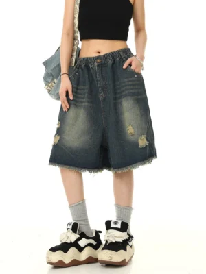 Front-left view of blue distressed denim Bermuda shorts styled with a black cropped tank top, chunky sneakers, grey socks, and a denim shoulder bag