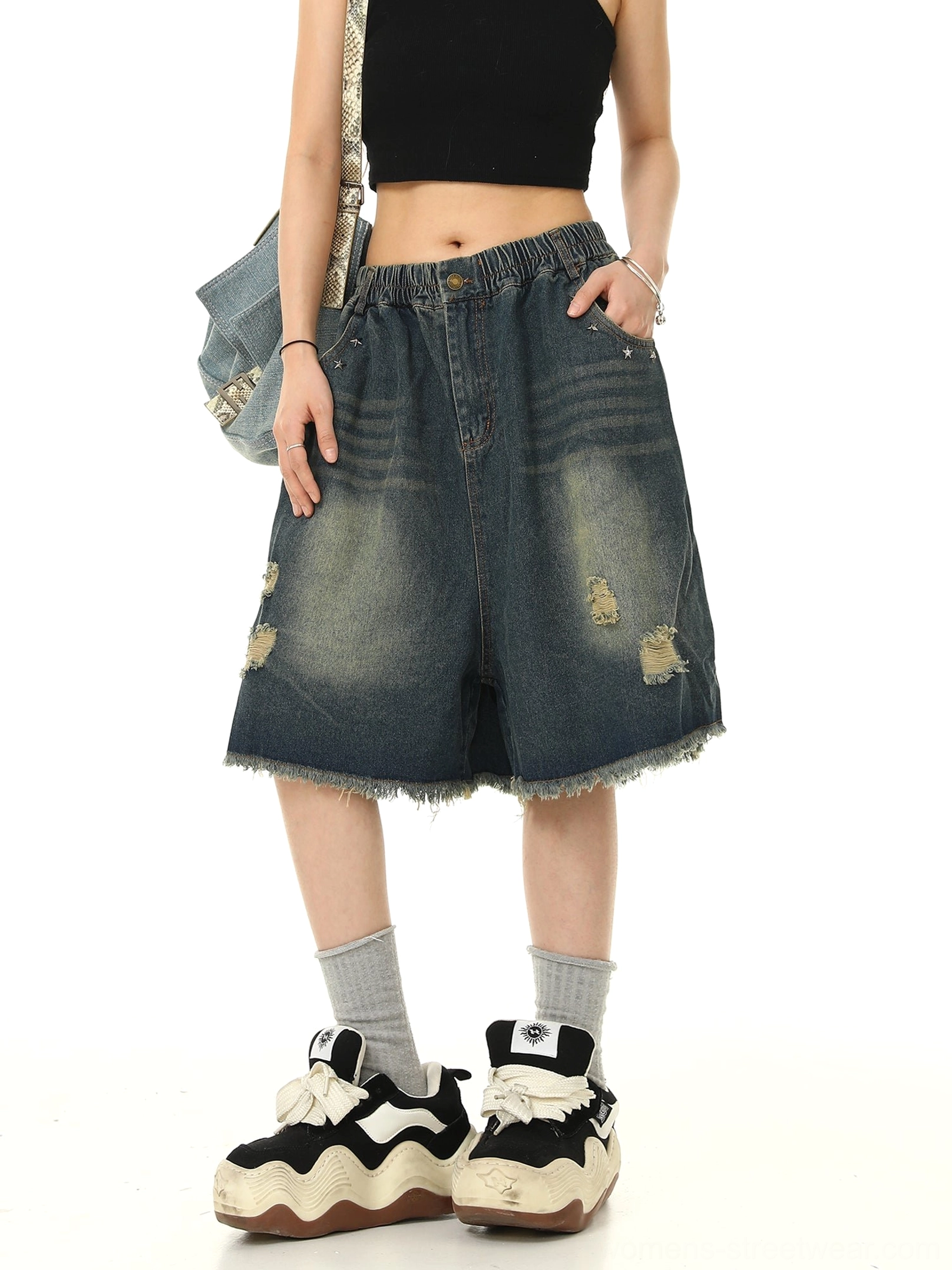 Front-left view of blue distressed denim Bermuda shorts styled with a black cropped tank top, chunky sneakers, grey socks, and a denim shoulder bag