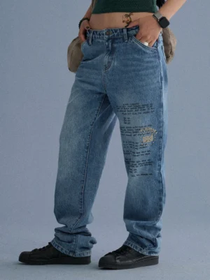Front view of high-waisted blue distressed jeans with thigh lettering, relaxed straight legs, crossbody bag, and skate shoes
