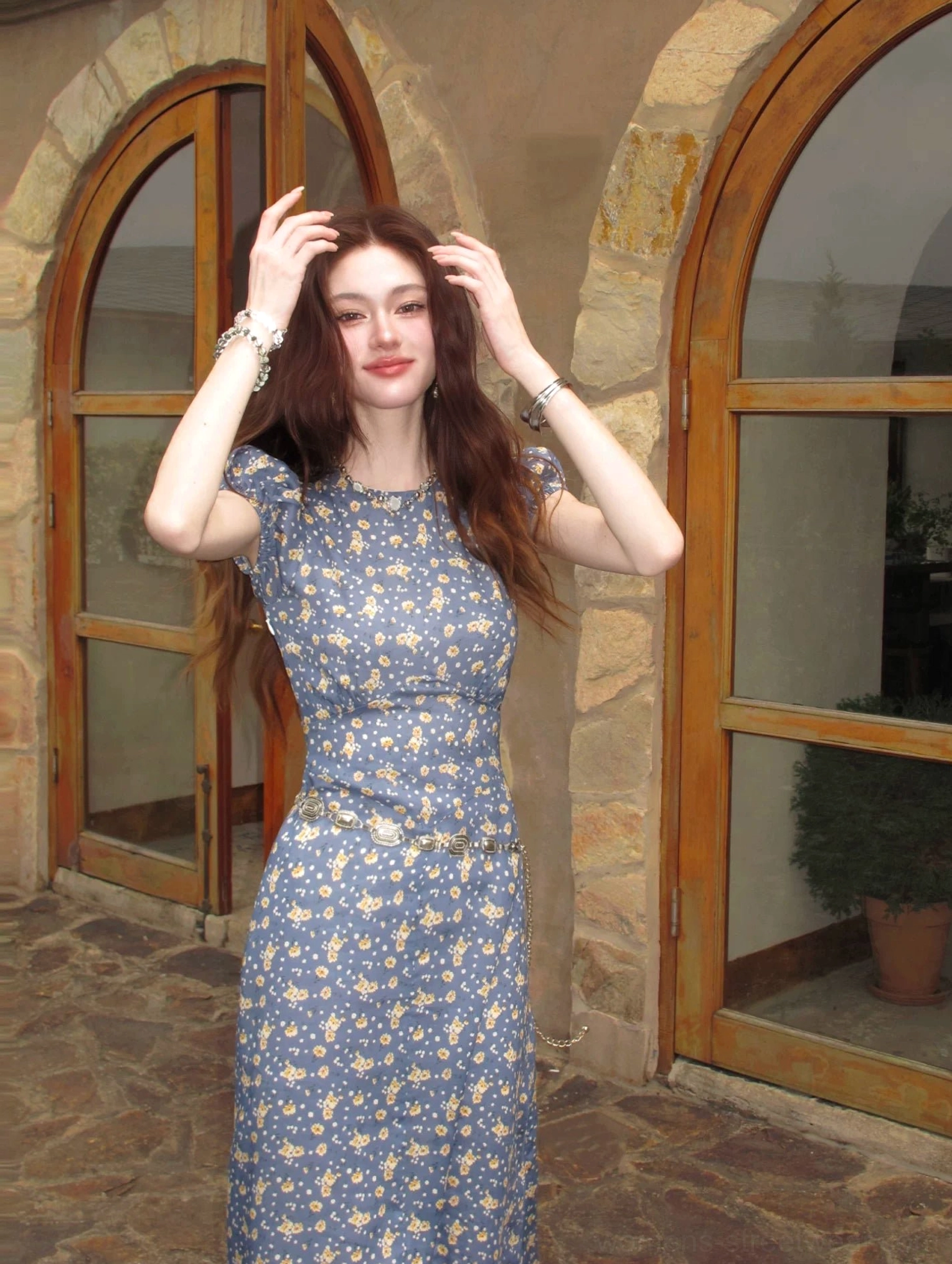 Blue ditsy floral open-back maxi dress with puff sleeves shown from the front in a stone villa courtyard