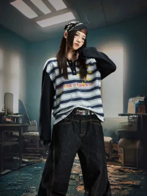 Front view of a female model wearing a blue grey striped rugby polo shirt with black contrast sleeves, graffiti lettering, and yellow star graphic in an abandoned retro office setting