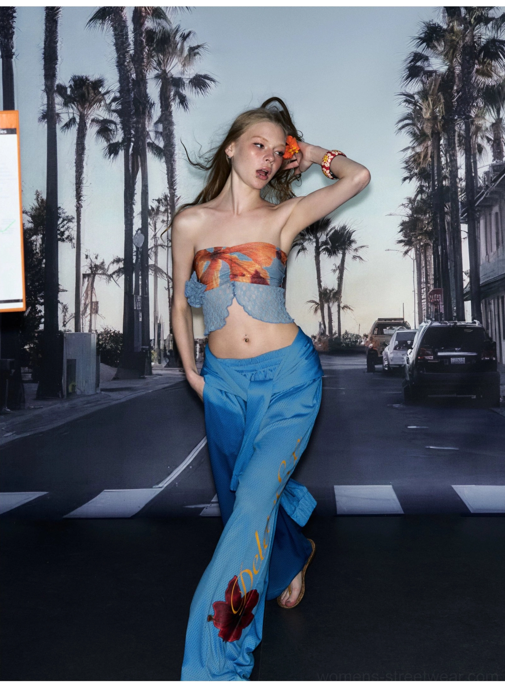 Blue high-waist hibiscus wide-leg streetwear pants styled in a dynamic side walking view at an urban crosswalk