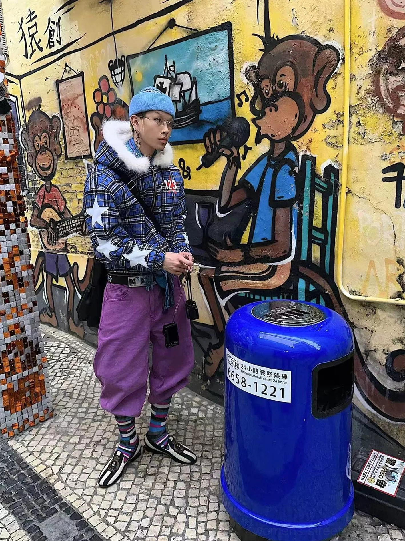 Blue plaid star puffer jacket with white graphic details and fur-trimmed hood worn with relaxed pants in a graffiti street setting