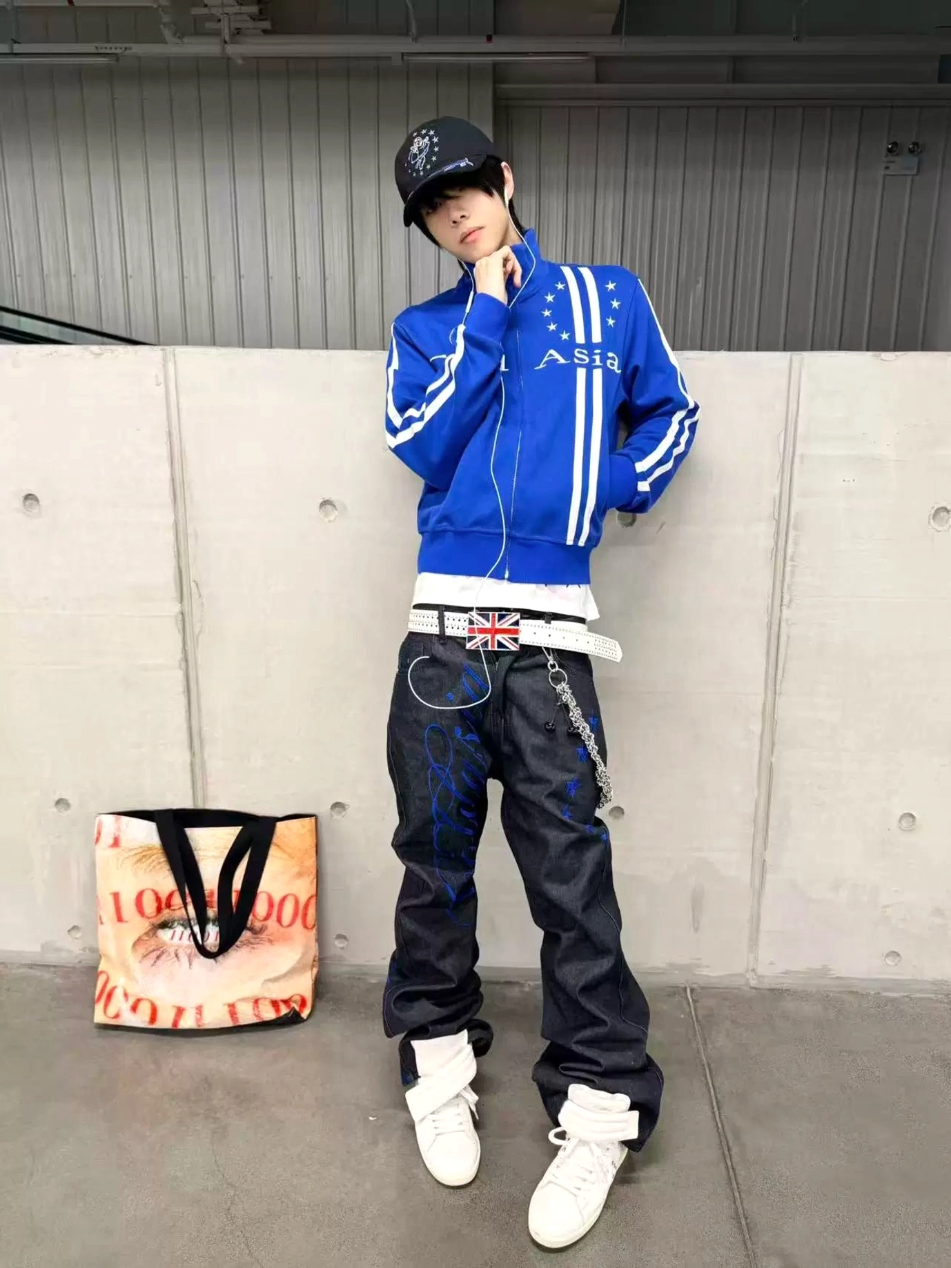 Blue retro track jacket styled with graphic jeans and streetwear accessories against a concrete wall, showing the sporty silhouette and white stripe details