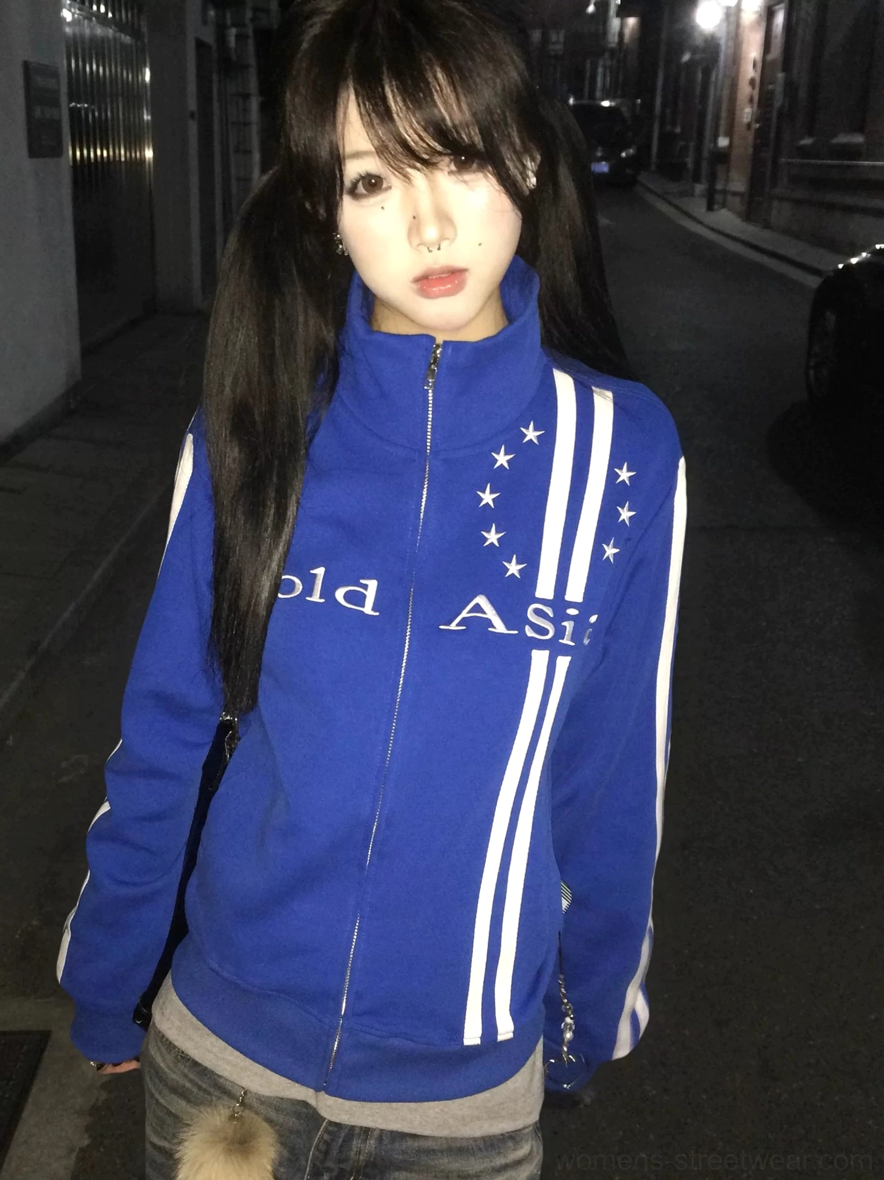 Royal blue starstripe track jacket worn by a female model in a nighttime urban alley, showing the stand collar full zip, white sleeve stripes, chest graphic, and retro Seoul streetwear style