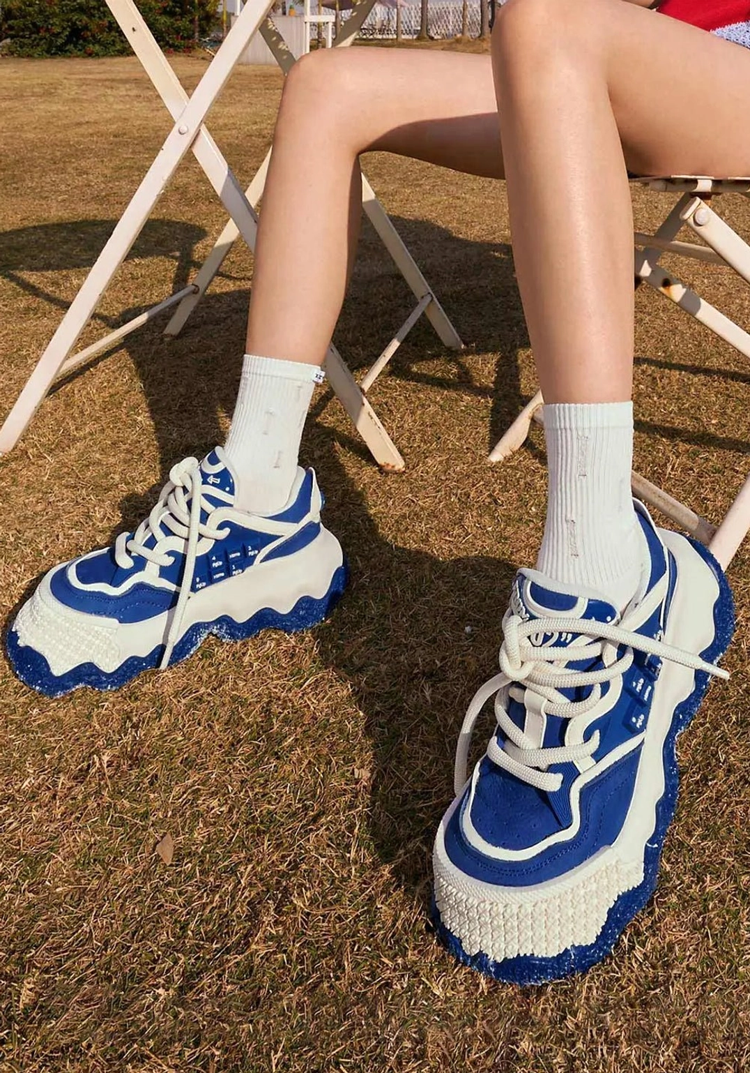 High angle view of female model wearing blue white chunky sole sneakers with white crew socks on dry grass, displaying the colorblock side panels and thick wavy soles