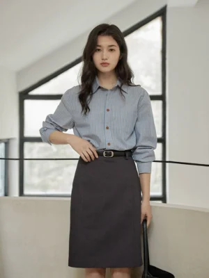 Full-body woman wearing a blue vertical striped shirt tucked into a tailored gray midi skirt with black belt and handbag