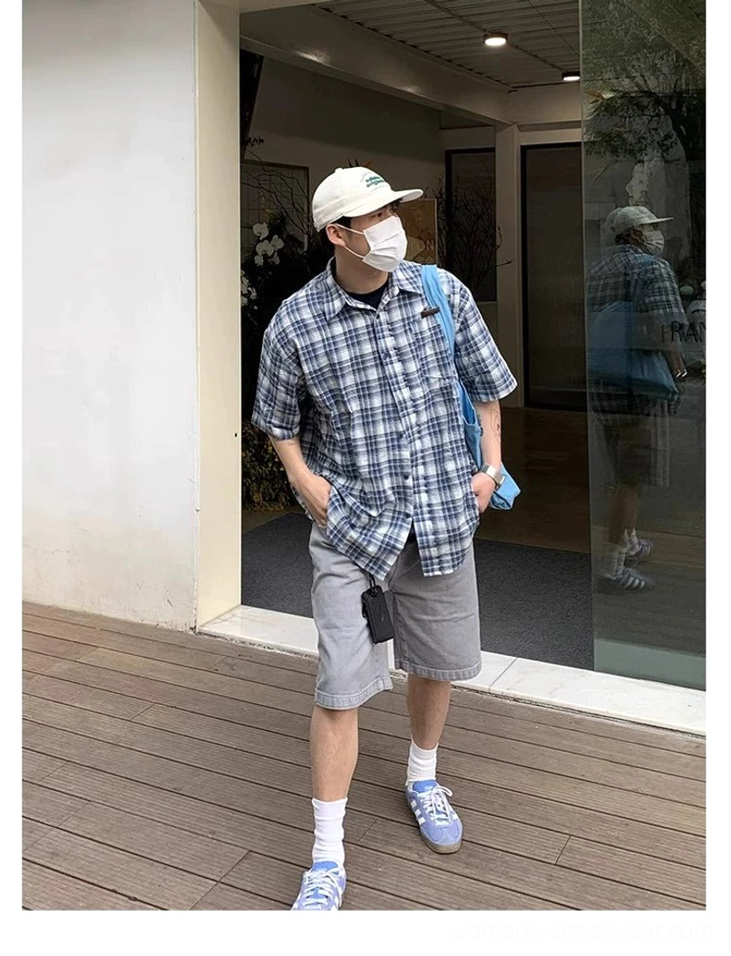 Side profile of a blue and white plaid oversized short sleeve shirt draped loosely over the torso and styled with relaxed shorts, cap, mask, and tote bag