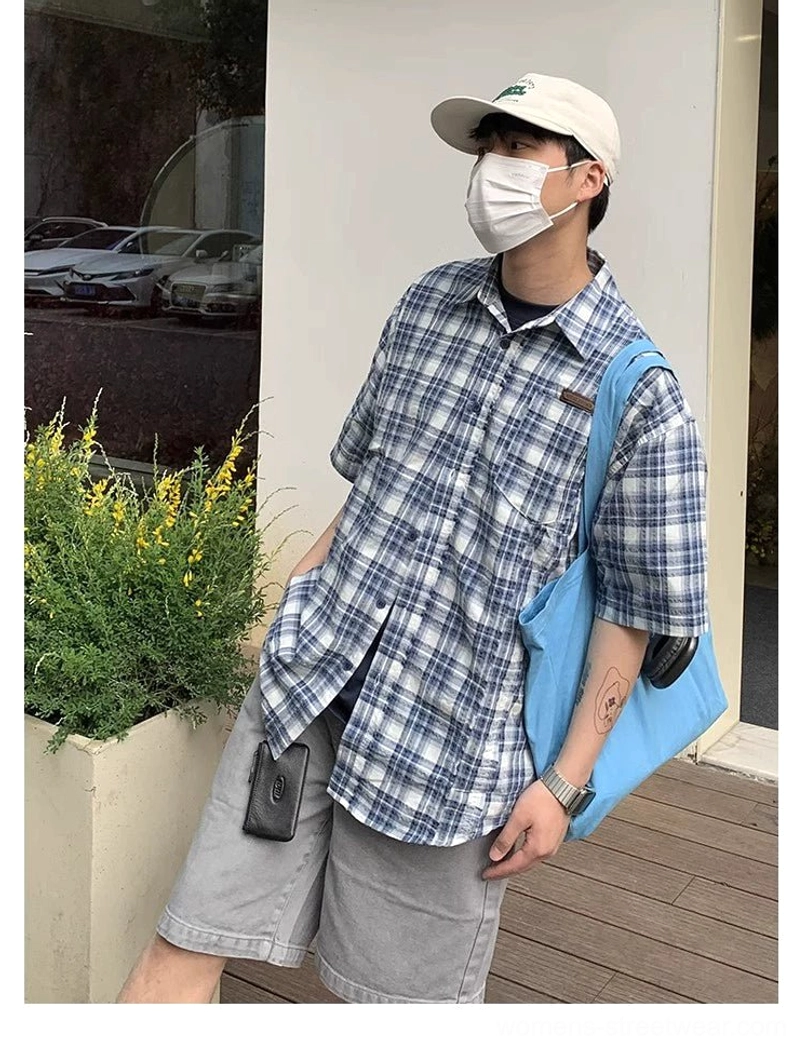 Three-quarter front view of a blue and white plaid short sleeve shirt worn partially open over a black T-shirt with a blue tote bag in a relaxed Korean fashion look