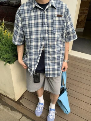 Close-up front view of a blue and white plaid short sleeve button-up shirt showing the classic grid pattern, button-down collar, chest patch pocket, and relaxed oversized fit