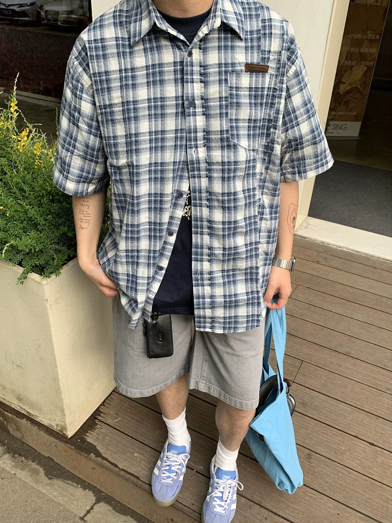 Close-up front view of a blue and white plaid short sleeve button-up shirt showing the classic grid pattern, button-down collar, chest patch pocket, and relaxed oversized fit