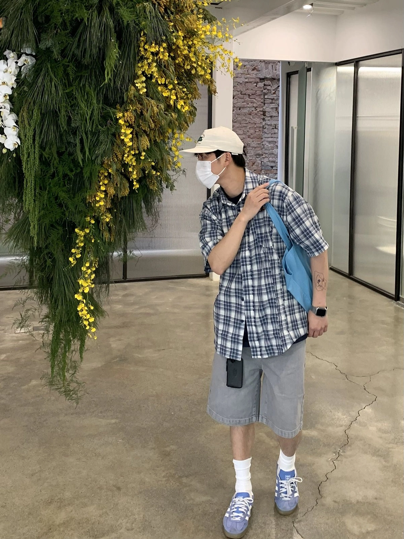 Side view of a relaxed oversized blue and white plaid short sleeve shirt worn partially unbuttoned over a black T-shirt with a blue tote bag for a casual layered streetwear look