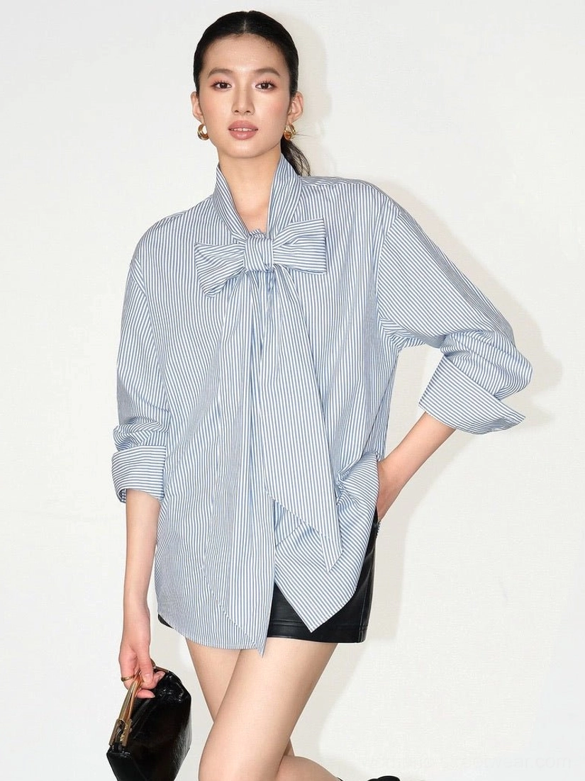 Asian female model wearing a blue and white striped oversized cotton bow blouse with rolled sleeves, black leather shorts, and a black clutch in a Korean streetwear outfit
