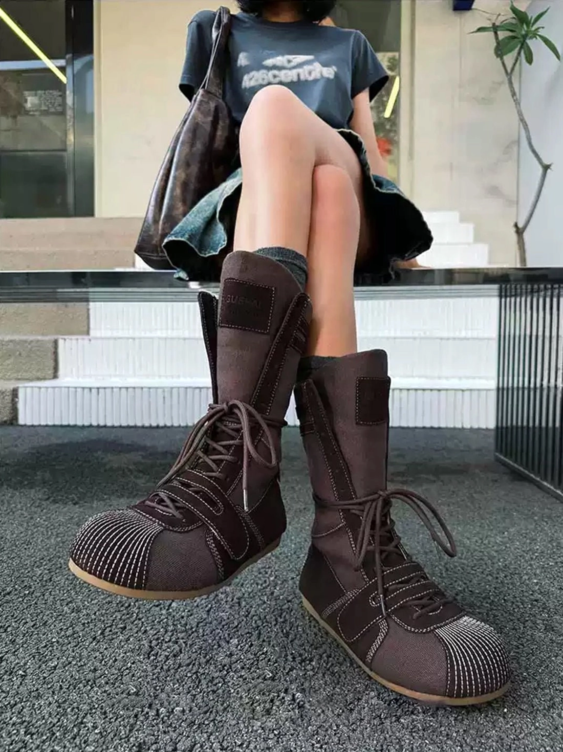 Three-quarter view of dark brown mid-calf boxing boots styled with a denim skirt and graphic tee on outdoor steps