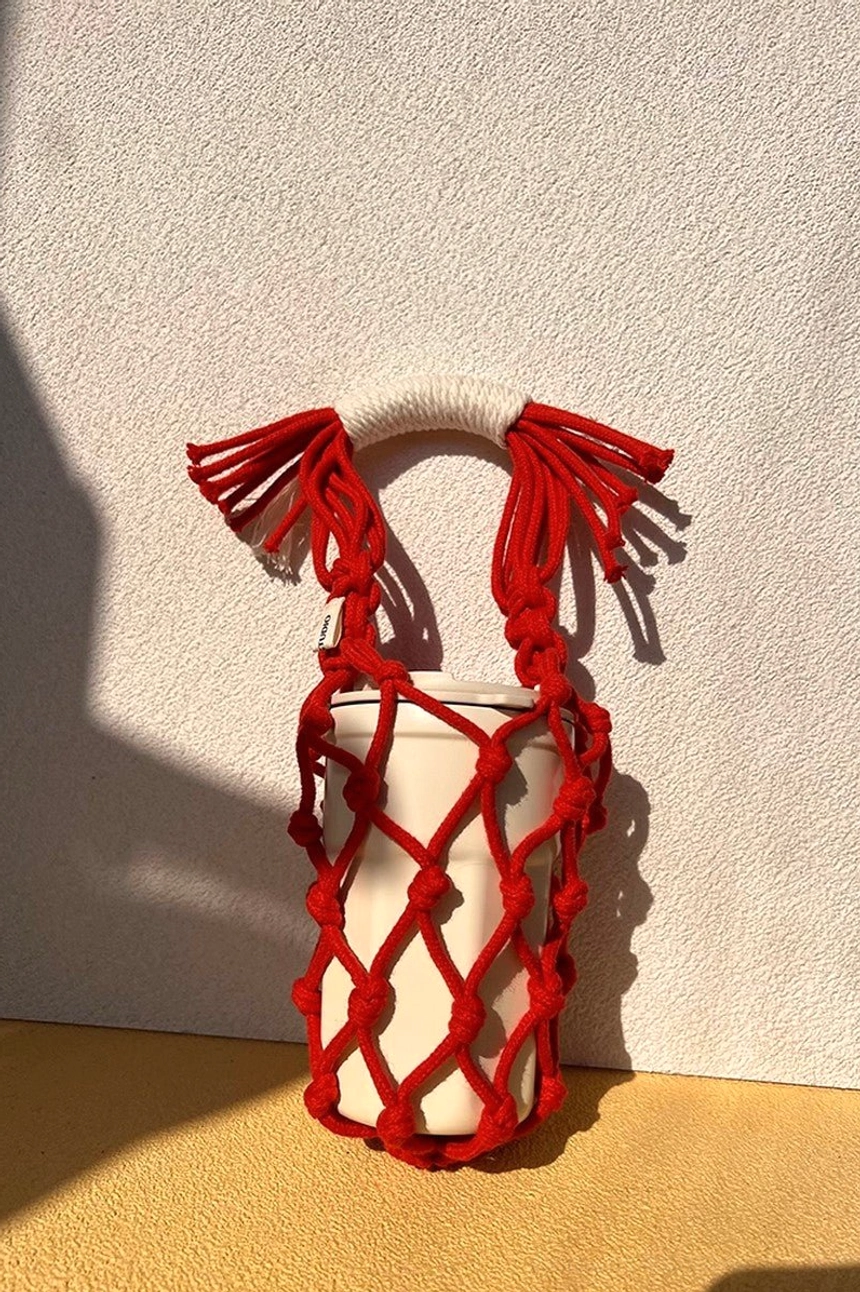 Bright red cotton macrame water bottle carrier holding a white tumbler, with textured woven rope construction and tassel handle