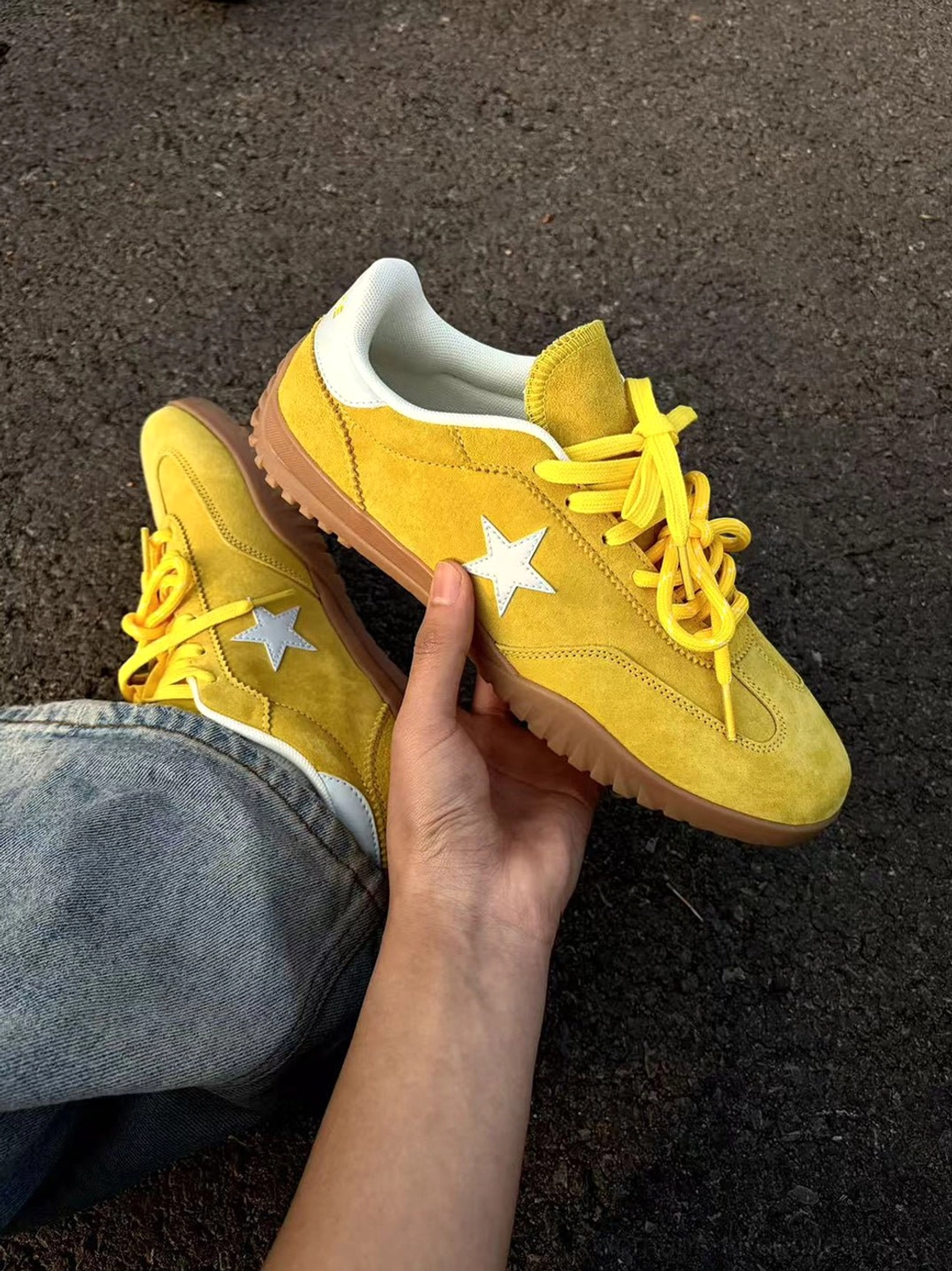 Handheld close-up of bright yellow suede star trainers with white emblem, lace-up closure, and white heel collar