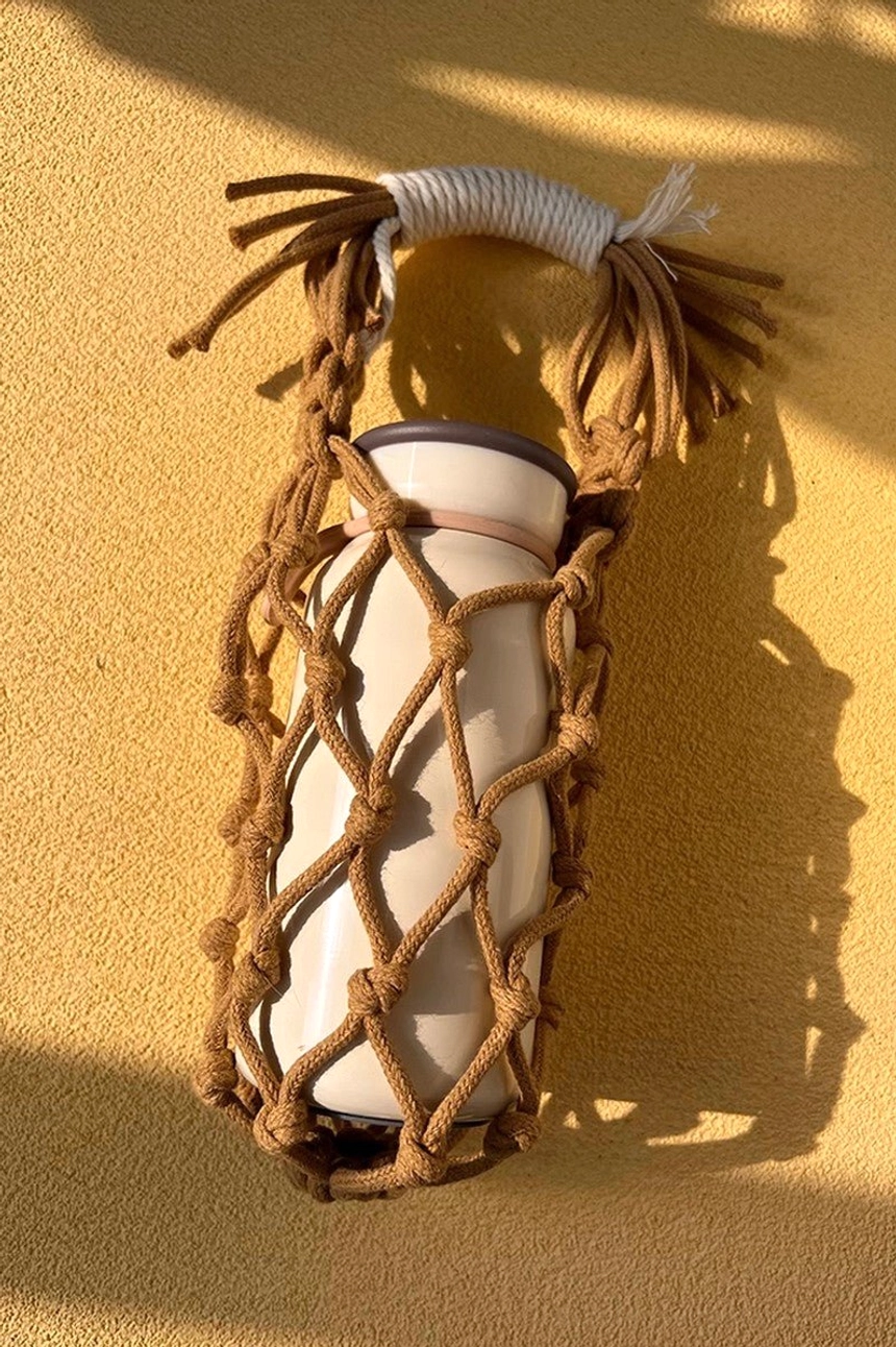 Brown cotton macrame water bottle carrier with white tumbler, showing the hand-knotted net pocket and wrapped tassel handle