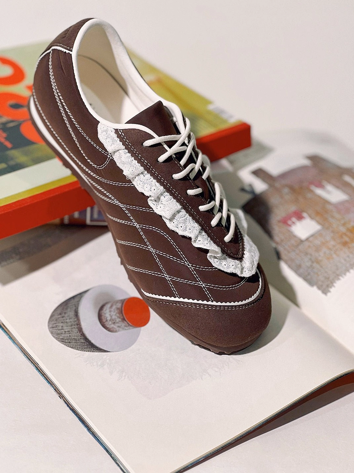 Brown frilled retro sneaker displayed on open books showing the white lace trim contrast stitching and low-profile sole