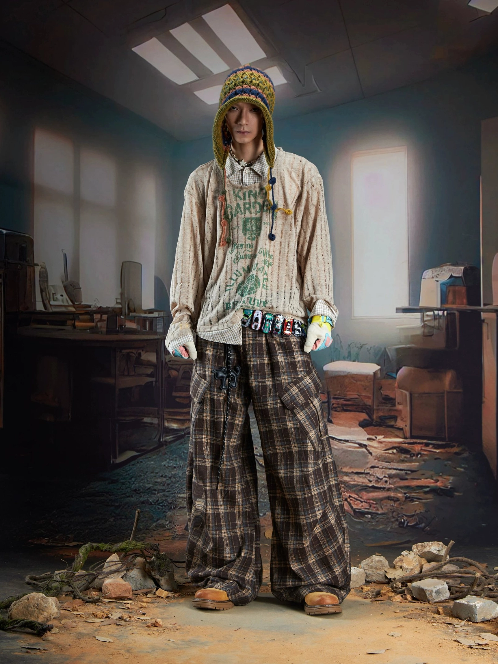 Male model styling a brown sheer ribbed layered vintage print sweater with green artwork, wide-leg plaid cargo pants, gloves, and decorative streetwear accessories