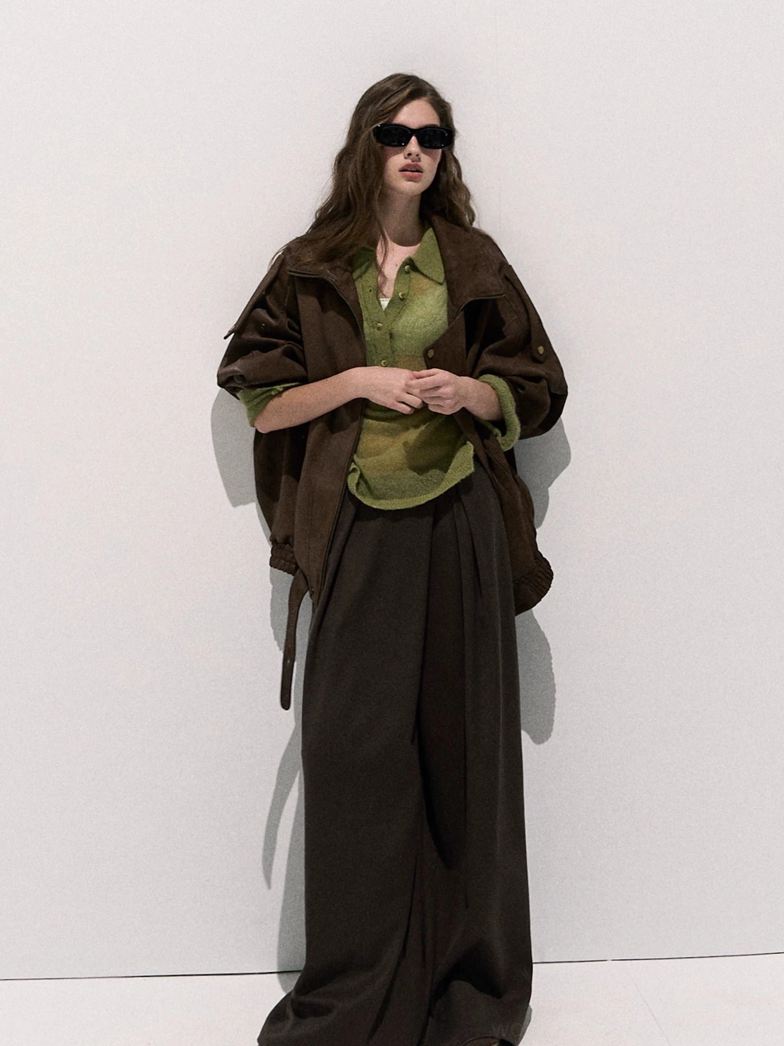 Full-body front view of a woman styling a brown oversized suede-look moto jacket with green shirt, wide-leg dark brown trousers, large lapel, and front zipper