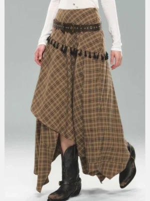 Close-up front view of brown plaid asymmetric skirt featuring black tassel trim, layered hem, and wide waist styling with black cowboy boots