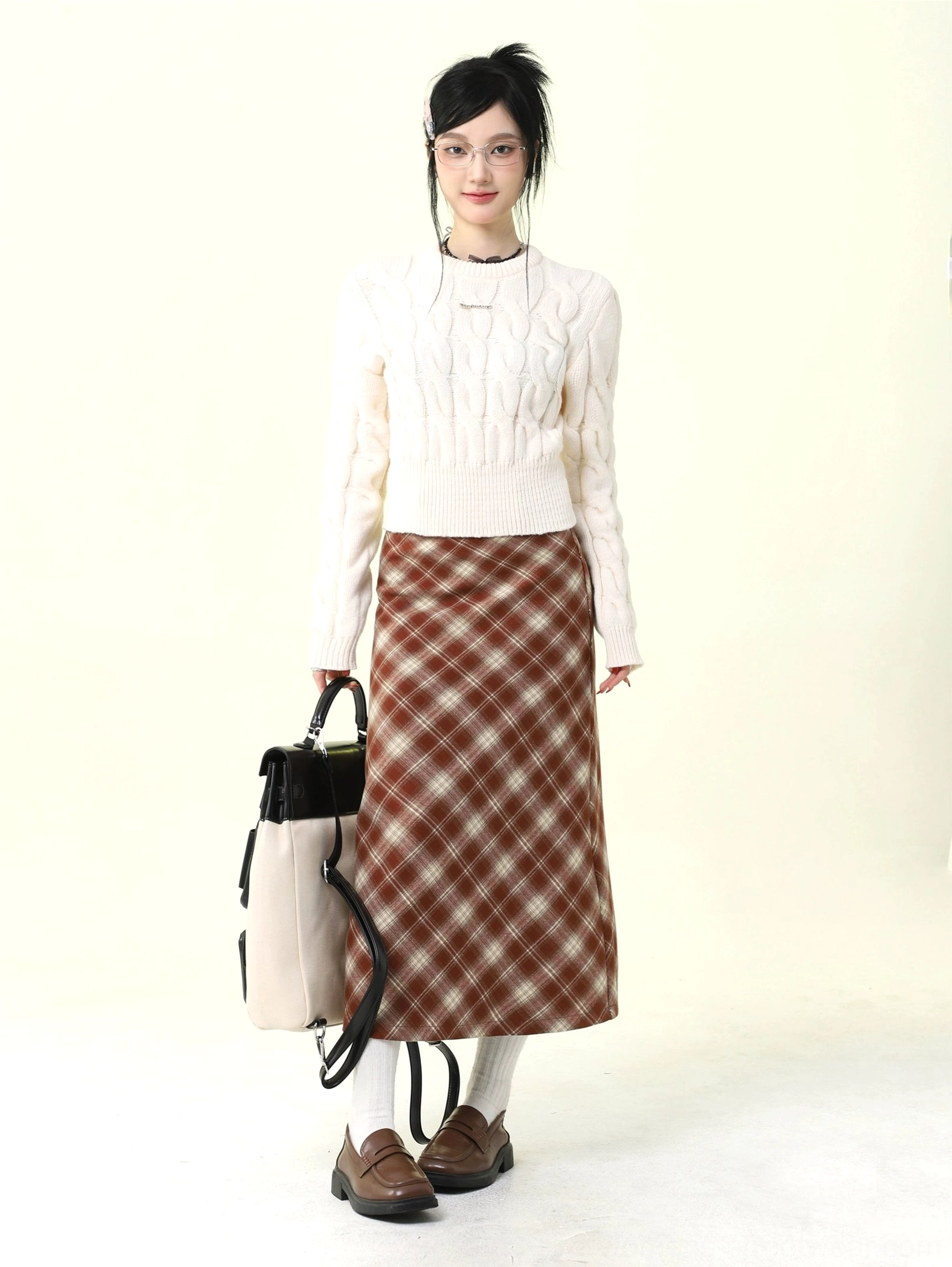 Full-body model wearing a warm brown red plaid wool skirt with a white cable-knit sweater and leather loafers