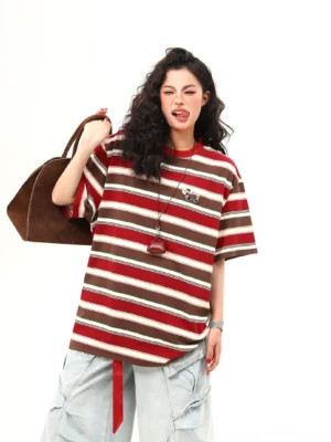 Front three-quarter view of a brown and red striped oversized streetwear T-shirt with embroidered chest detail, red hem strap, wide-leg denim shorts, and brown leather tote