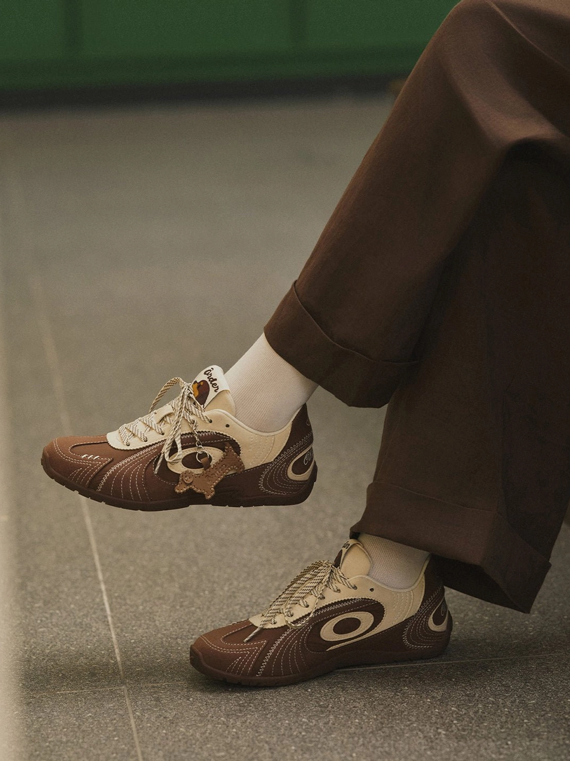 Side view of brown retro trainers with cream swirl overlays and dog-shaped charm worn with cropped brown trousers