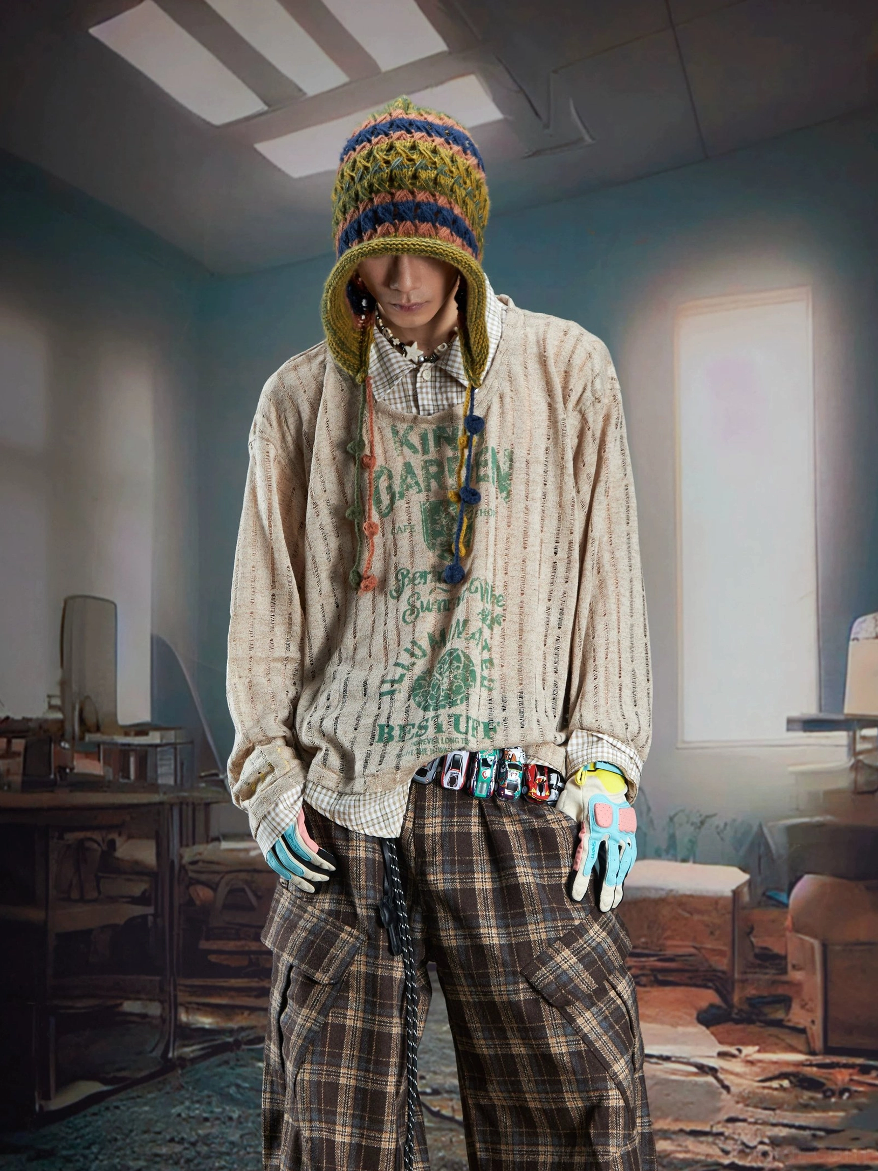 Male model wearing a brown ribbed sheer layered streetwear sweater with green retro print, colorful gloves, decorative belt, and relaxed plaid cargo pants