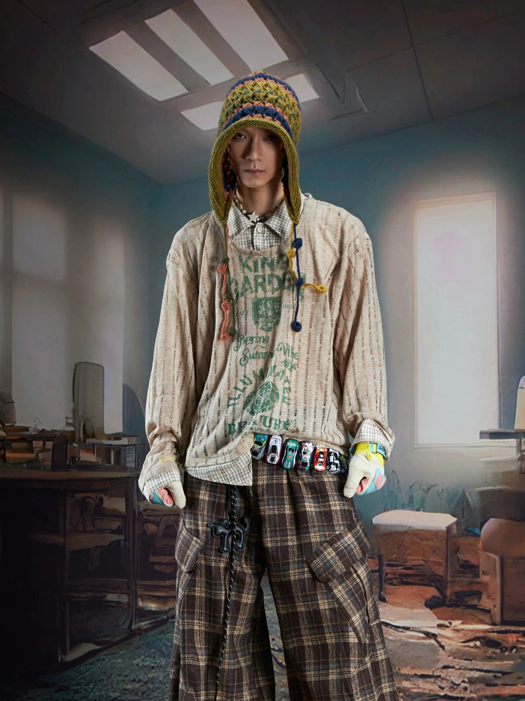 Low angle view of a male model in a brown sheer layered vintage graphic sweater with green print, plaid cargo pants, and hanging chain accessory