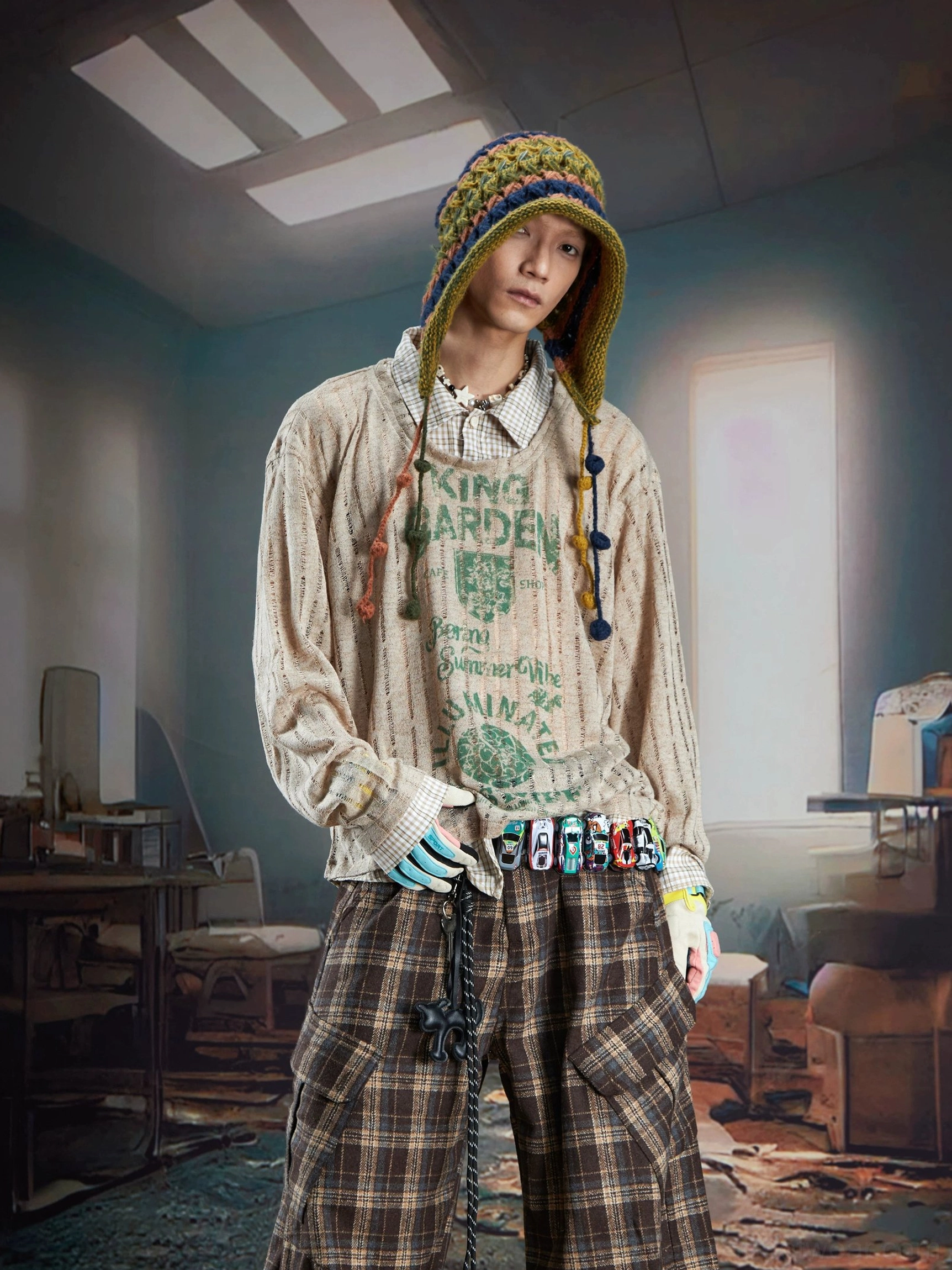 Male model wearing a brown sheer ribbed layered shirt sweater with green vintage graphics, crochet hat, colorful belt, gloves, and loose cargo pants