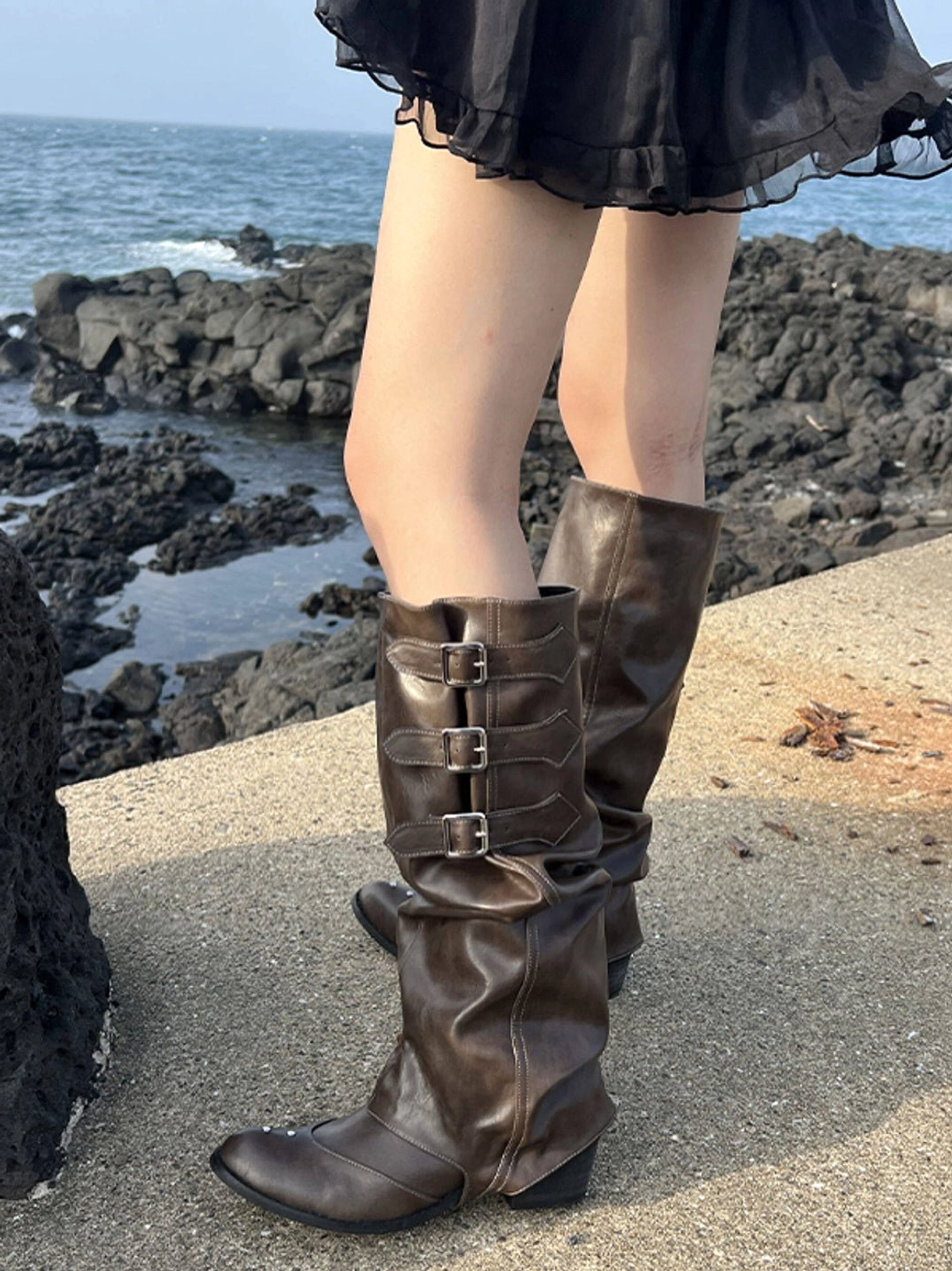 Brown slouchy western knee-high boots with triple buckles styled with a black mini skirt in an outdoor streetwear look