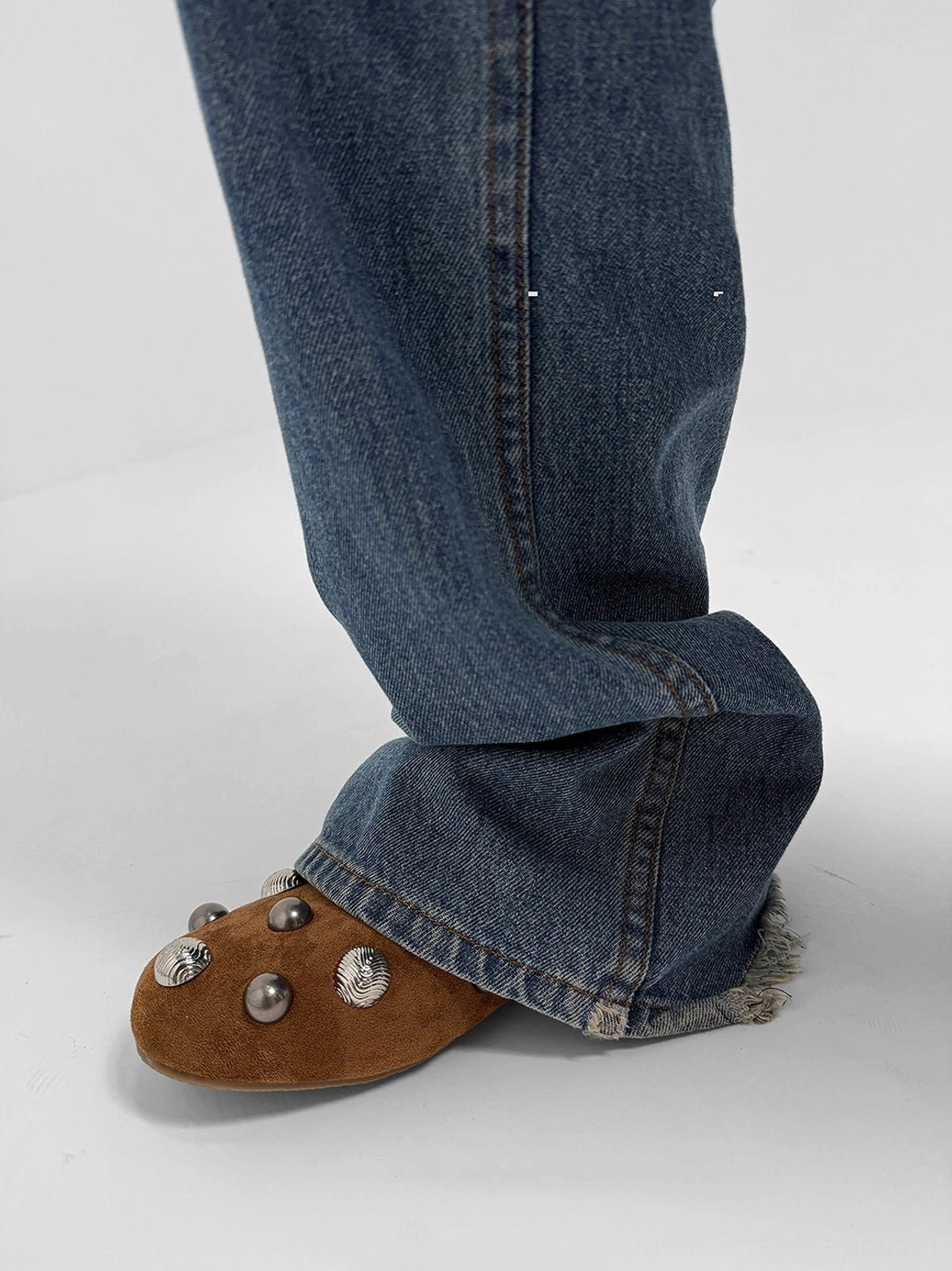 Brown suede-look ballet flat with decorative studs worn with blue jeans, showing the slim flat sole and low-profile shape