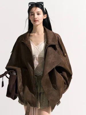 Front view of a woman wearing a brown suede-look oversized moto jacket with wide lapels, shoulder snap buttons, adjustable drawstring hem, lace camisole, and embroidered mini skirt