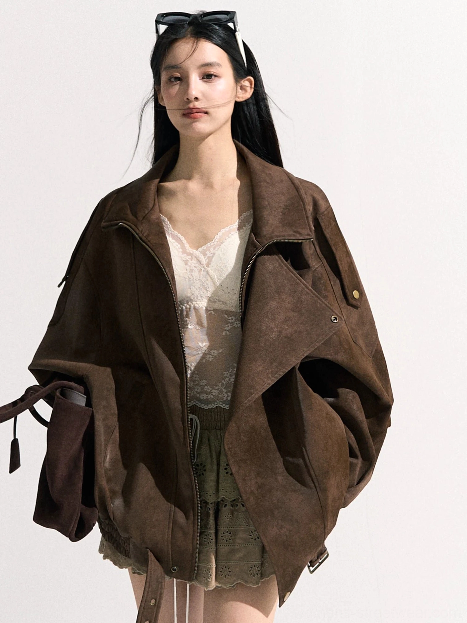 Front view of a woman wearing a brown suede-look oversized moto jacket with wide lapels, shoulder snap buttons, adjustable drawstring hem, lace camisole, and embroidered mini skirt